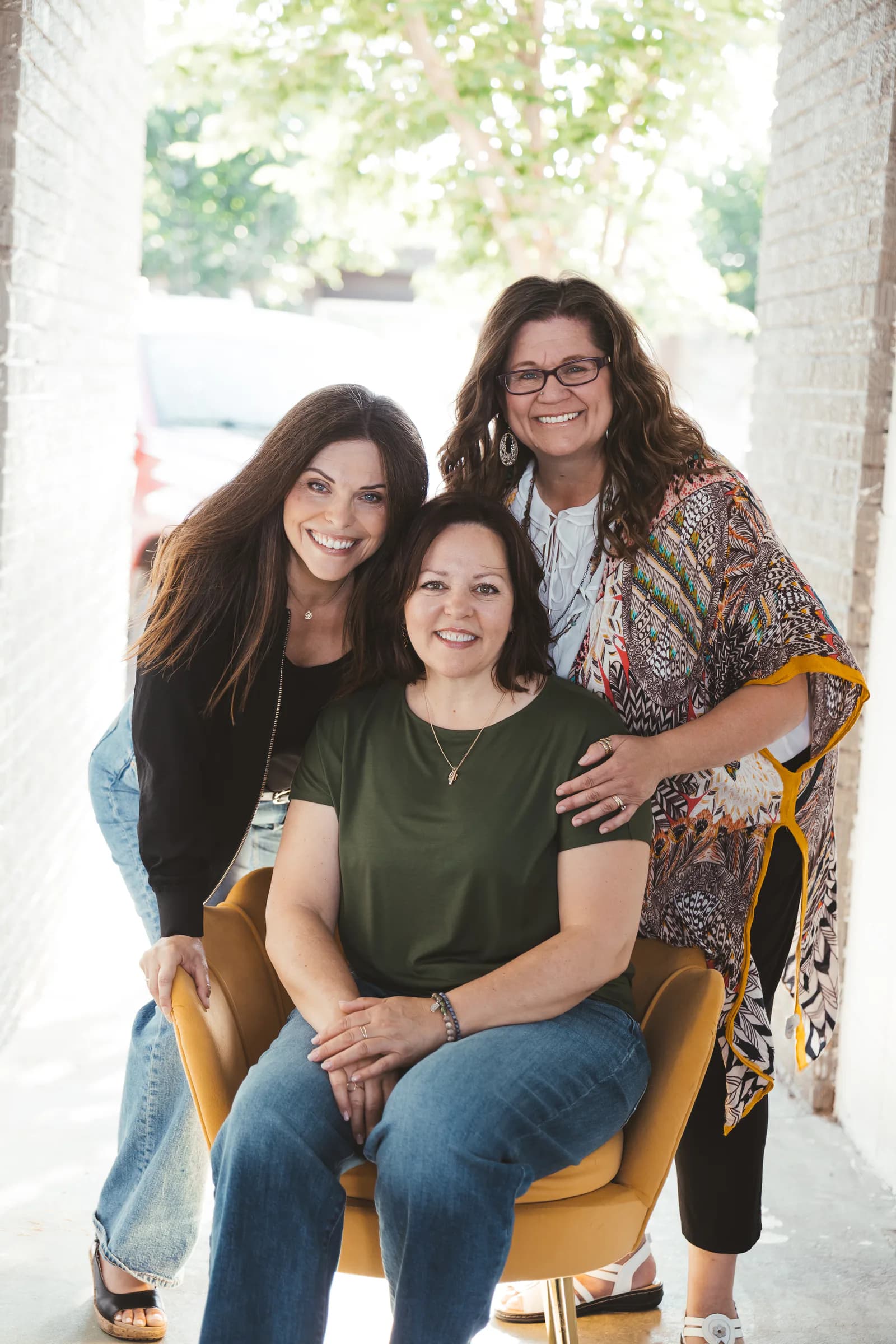 Kelly, Deena and Krista smiling together outdoors, with one seated in a yellow chair and two standing close behind her.